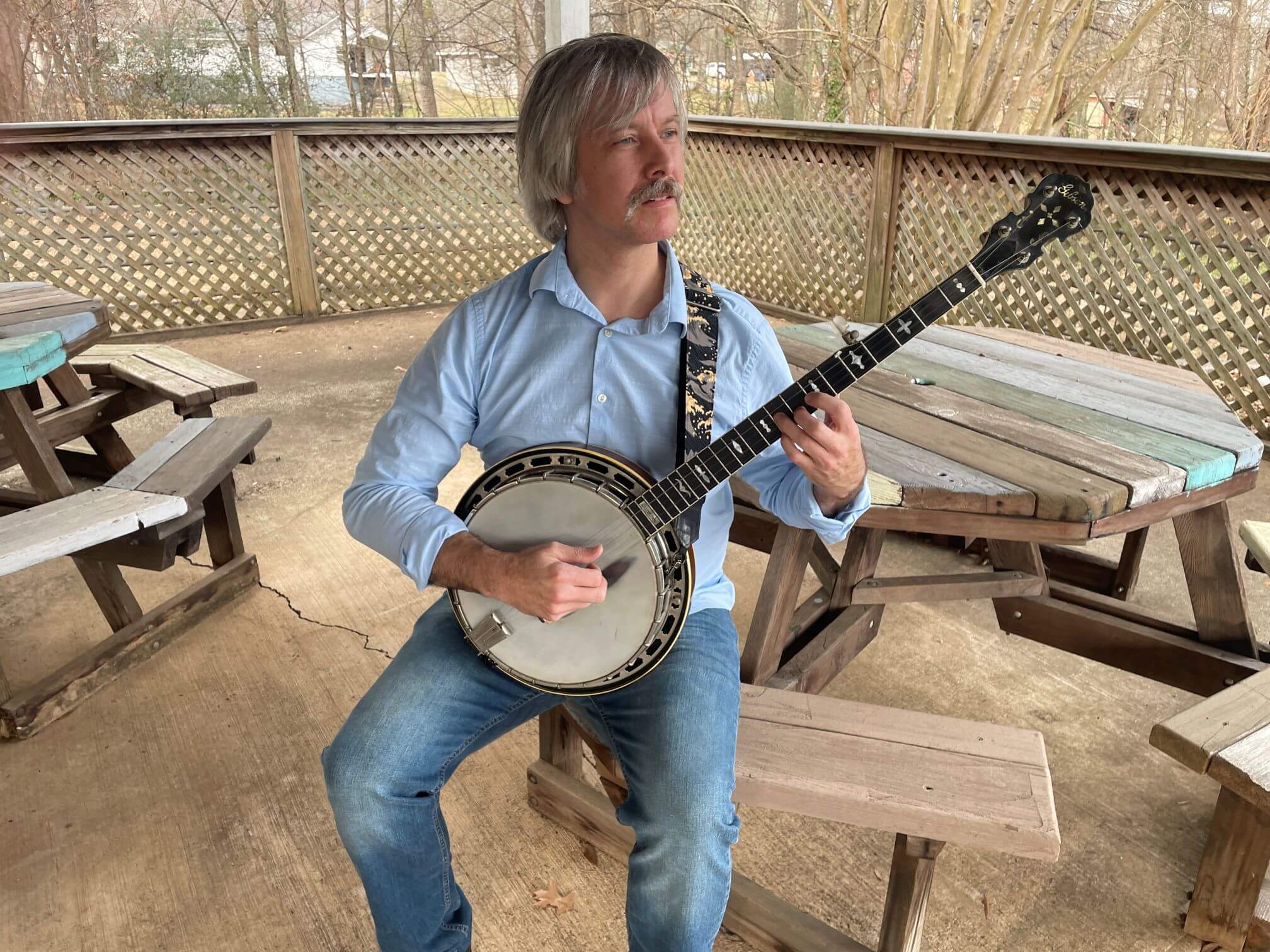 man sitting on a wooden bench playing banjo in a blue shirt